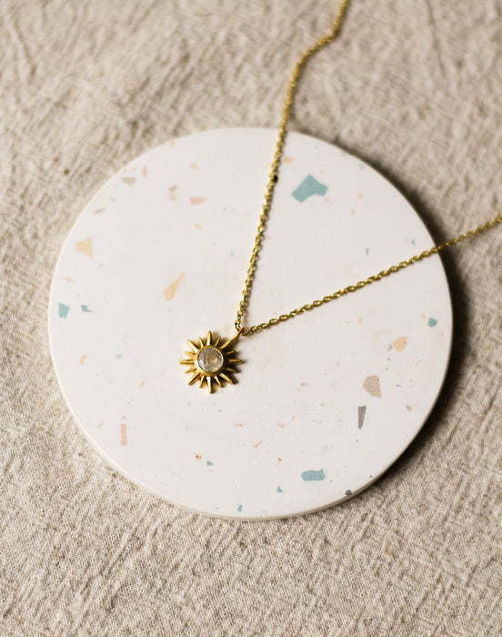 Gold necklace with a sun pendant on a round, speckled coaster on a beige linen surface