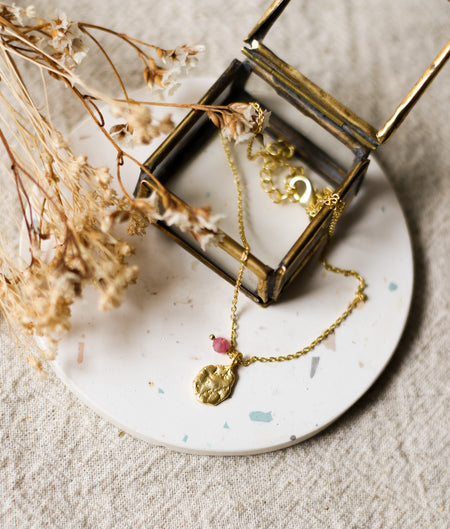 Gold necklace with a pendant on a speckled white coaster with dried flowers and a glass jewellery box