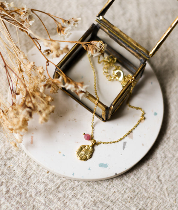 Gold necklace with a pendant on a speckled white coaster with dried flowers and a glass jewellery box