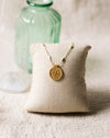 Gold necklace with a gold disc pendant on a beige pillow with a green glass bottle in the background on a beige linen surface