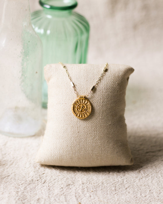 Gold necklace with a gold disc pendant on a beige pillow with a green glass bottle in the background on a beige linen surface