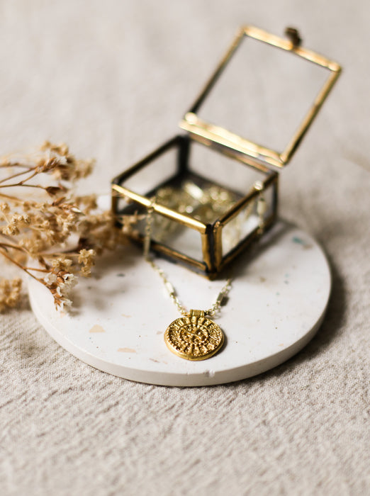 Gold necklace with a gold disc pendant on a white speckled coaster next to a glass jewellery box and dried flowers