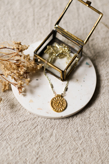 Gold necklace with a gold disc pendant on a white speckled coaster next to a glass jewellery box and dried flowers