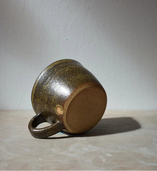 dark green ceramic mug laying on its side, on a light surface with a grey background