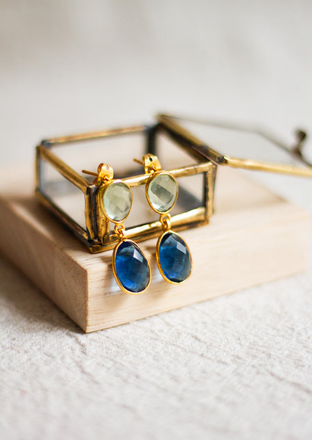 Gold earrings with blue and green gemstones on a wooden stand