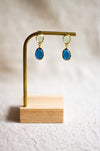 Pair of gold and blu earrings on a brass and wooden stand against a neutral linen background