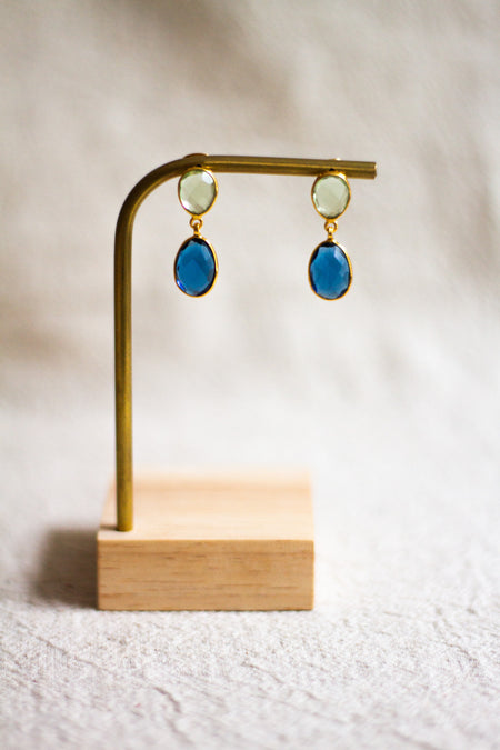 Pair of gold and blu earrings on a brass and wooden stand against a neutral linen background