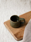 Green ceramic espresso cup on a wooden chopping board with a striped tea towel