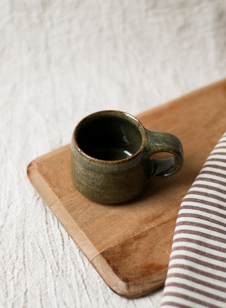 Green ceramic espresso cup on a wooden chopping board with a striped tea towel