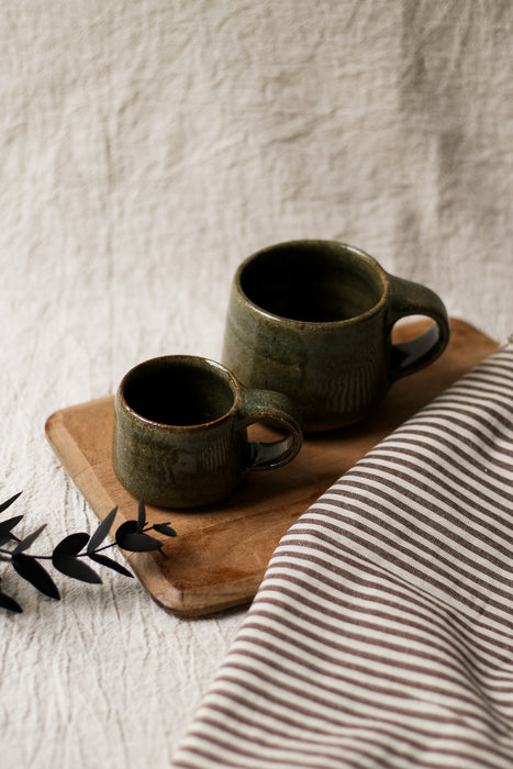 Two dark green ceramic mugs on a wooden chopping board with a striped tea towel and eucalyptus in the background.