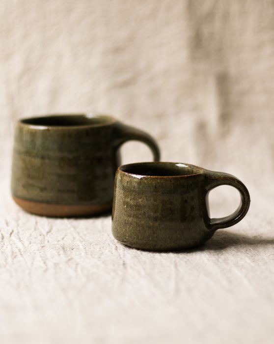 Two dark green ceramic mugs on a beige   linen surface