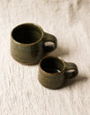 Two dark green ceramic mugs on a beige linen surface