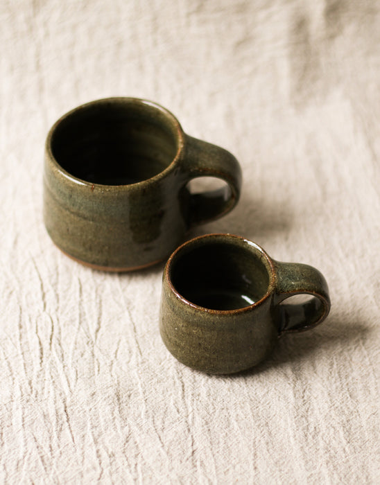 Two dark green ceramic mugs on a beige linen surface