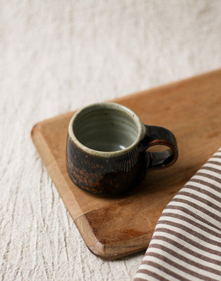 Brown ceramic espresso cup on a wooden bread board with a striped cloth in the background