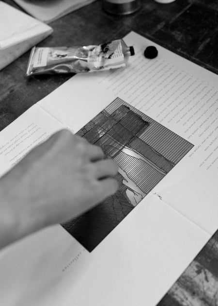 Artists hand working on an etching on a printing plate with a tube of ink laying next to it on a wooden table 