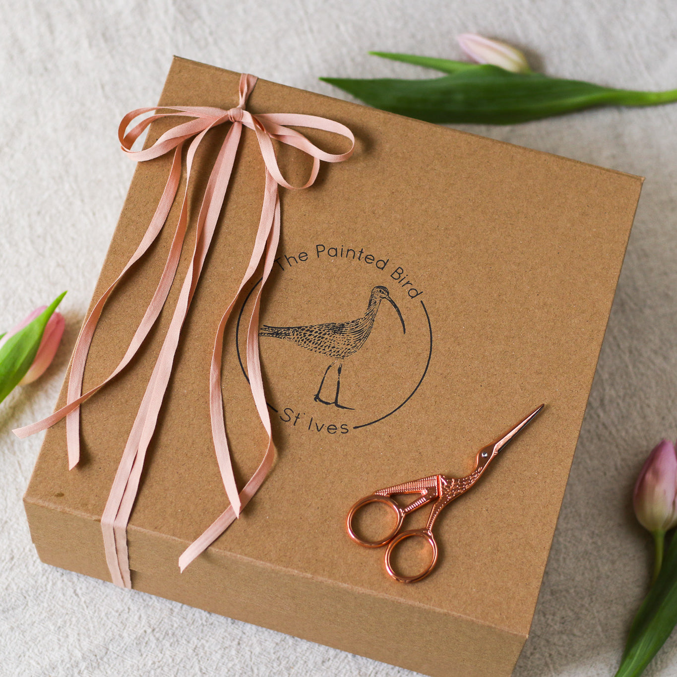 Brown paper gift box with The Painted Bird logo, sewing scissors on top, tied with pink ribbon, and surrounded with pink tulips, on a beige linen surface 