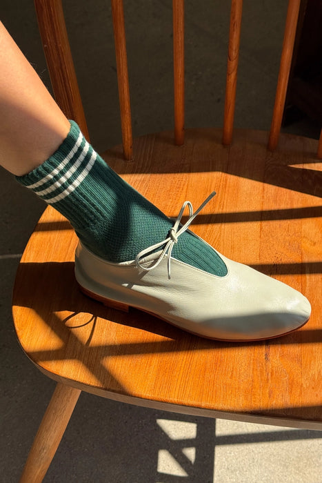 Person wearing green socks with white stripes and sage flat shoes on a wooden chair.