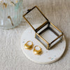 Two gold hoop earrings displayed on a small round white coaster with a glass and brass jewellery box, next to a glass vase with dried flowers on a textured beige fabric background.