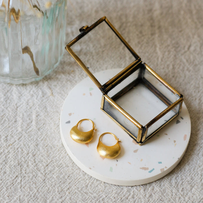 Two gold hoop earrings displayed on a small round white coaster with a glass and brass jewellery box, next to a glass vase with dried flowers on a textured beige fabric background.
