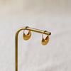 A pair of small gold crescent-shaped hoop earrings hanging from a brass display stand against a soft light beige background.