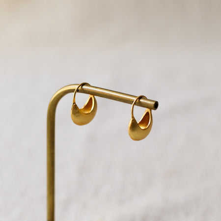 A pair of small gold crescent-shaped hoop earrings hanging from a brass display stand against a soft light beige background.