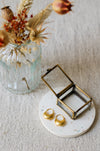Two gold hoop earrings displayed on a round white coaster next to an brass jewellery box, with a glass vase of dried flowers in the background on a textured light linen surface.