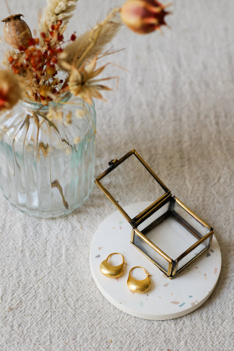 Two gold hoop earrings displayed on a round white coaster next to an brass jewellery box, with a glass vase of dried flowers in the background on a textured light linen surface.