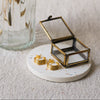A pair of gold hoop earrings displayed on a round white coaster next to a small open glass jewellery box, with a ribbed glass vase in the background.