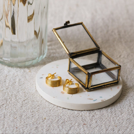 A pair of gold hoop earrings displayed on a round white coaster next to a small open glass jewellery box, with a ribbed glass vase in the background.