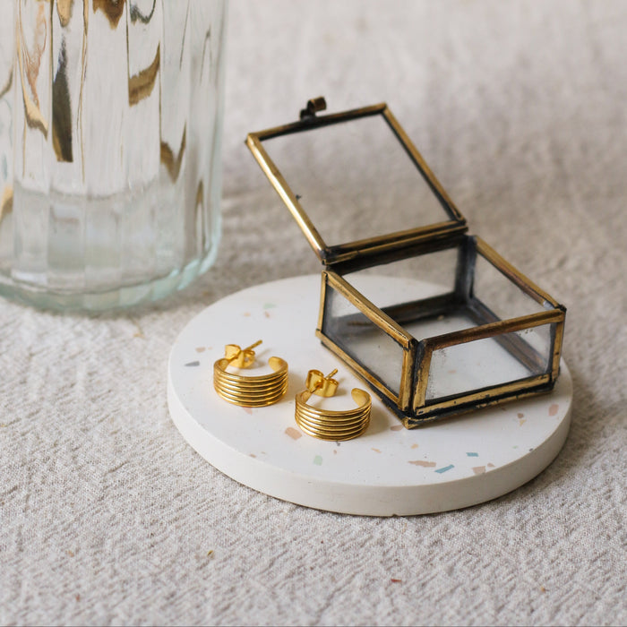 A pair of gold hoop earrings displayed on a round white coaster next to a small open glass jewellery box, with a ribbed glass vase in the background.