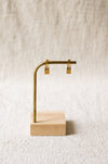 A pair gold hoop earrings displayed on a brass stand with a light wood base against a textured beige fabric background.