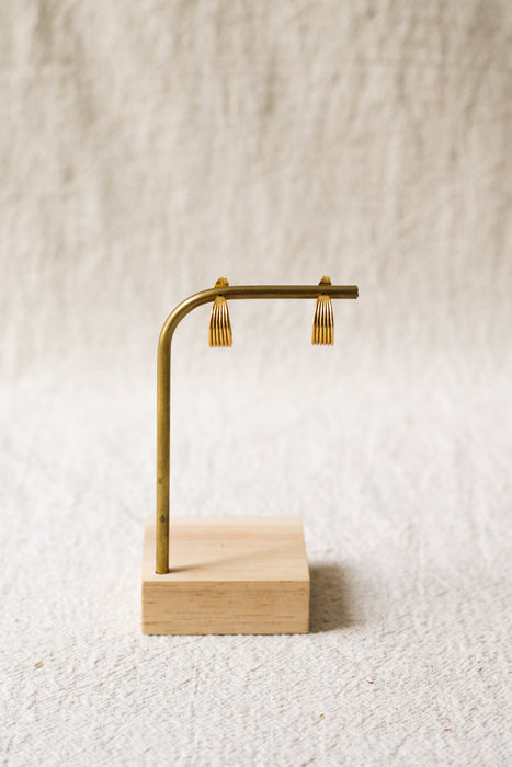 A pair gold hoop earrings displayed on a brass stand with a light wood base against a textured beige fabric background.