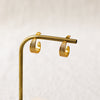 A pair of gold hoop earrings with a textured, ridged design, displayed on a brass stand against a soft, light beige background.