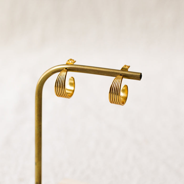 A pair of gold hoop earrings with a textured, ridged design, displayed on a brass stand against a soft, light beige background.