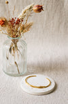 A thin gold bangle bracelet displayed on a round white coaster, with a ribbed clear glass vase holding dried flowers in the background against a textured beige fabric backdrop.