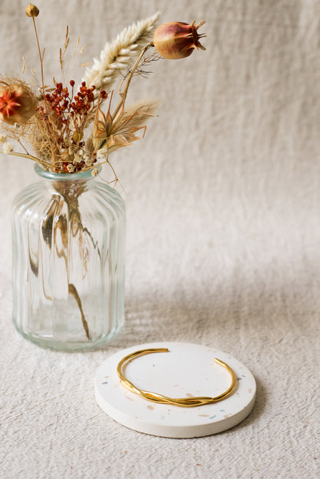A thin gold bangle bracelet displayed on a round white coaster, with a ribbed clear glass vase holding dried flowers in the background against a textured beige fabric backdrop.