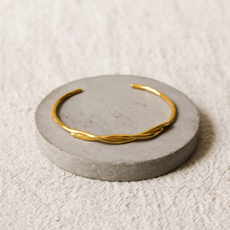 A thin gold twisted cuff bracelet displayed on a round grey concrete riser over a textured beige linen background.
