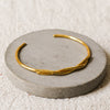 A thin gold twisted cuff bracelet displayed on a round grey concrete riser over a textured beige linen background.