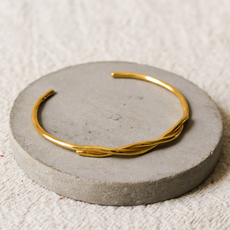 A thin gold twisted cuff bracelet displayed on a round grey concrete riser over a textured beige linen background.