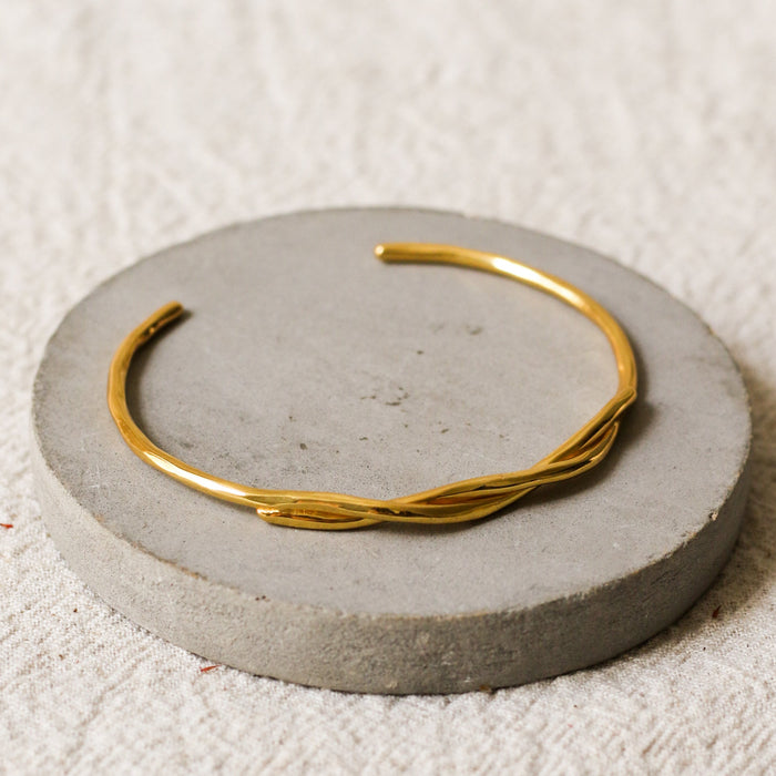 A thin gold twisted cuff bracelet displayed on a round grey concrete riser over a textured beige linen background.