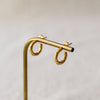 A pair of textured gold hoop earrings displayed on a brass stand with a light wood base against a soft, neutral background.