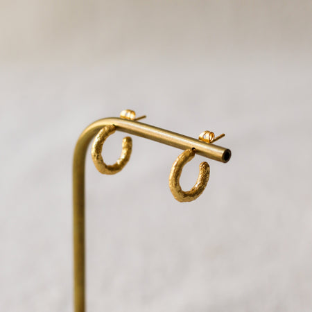 A pair of textured gold hoop earrings displayed on a brass stand with a light wood base against a soft, neutral background.