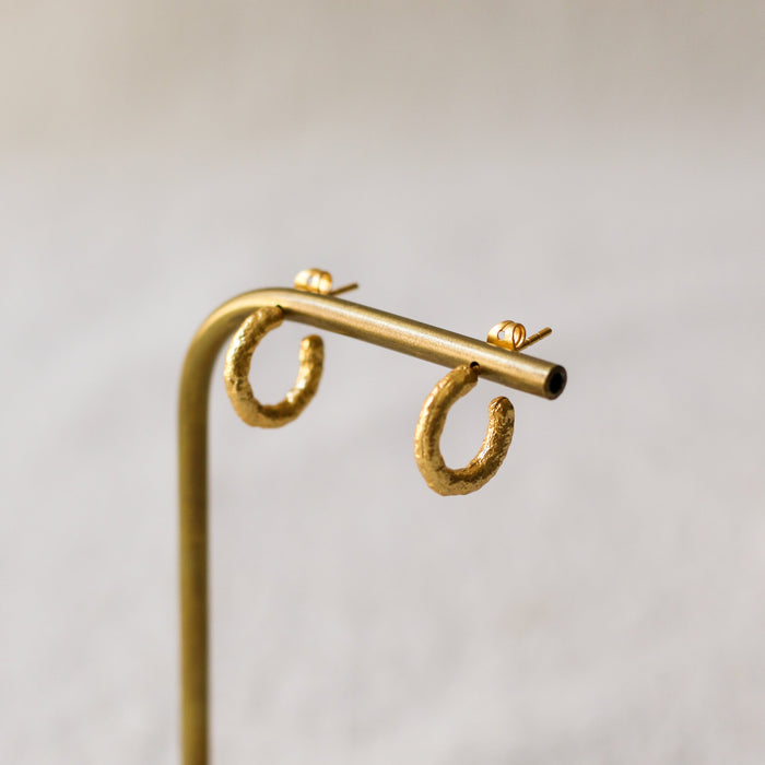 A pair of textured gold hoop earrings displayed on a brass stand with a light wood base against a soft, neutral background.
