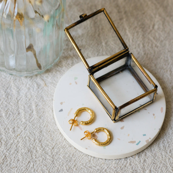 Gold hoop earrings displayed on a round white coaster next to an open brass jewellery box, with a glass vase of dried flowers in the background on a textured beige linen surface.