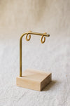 A pair of textured gold hoop earrings displayed on a brass stand with a light wood base against a soft, neutral background.