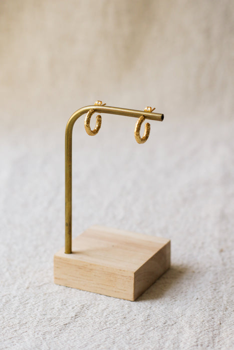 A pair of textured gold hoop earrings displayed on a brass stand with a light wood base against a soft, neutral background.