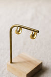 A pair of textured gold hoop earrings hanging from a brass display stand with a light wood base, set against a soft, out-of-focus neutral background.
