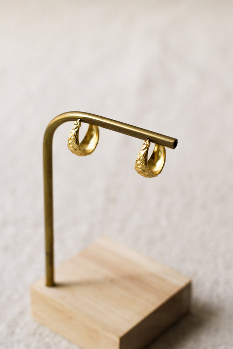 A pair of textured gold hoop earrings hanging from a brass display stand with a light wood base, set against a soft, out-of-focus neutral background.