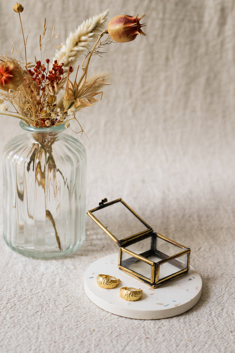 A small open brass jewellery box with gold hoop earrings sit on a round white coaster beside a glass vase holding dried flowers, all set on a textured beige fabric background.