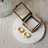 A small open jewelry box with a brass metal frame and clear glass lid sits on a round white speckled coaster. Two gold hoop earrings rest on the tray beside the box.
